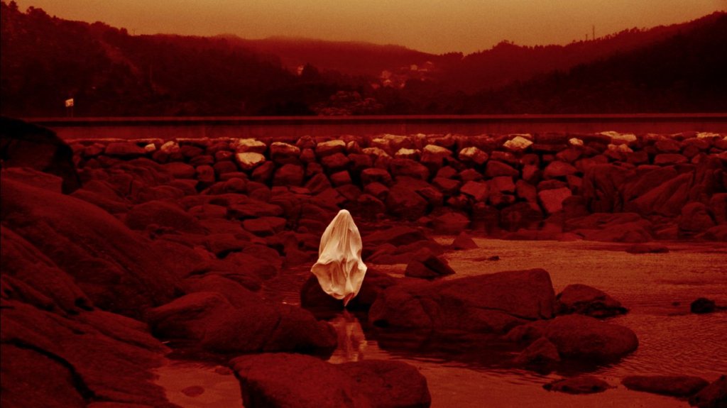 A shrouded figure sitting on the stones on a shore. Everything is drenched in a red light.
