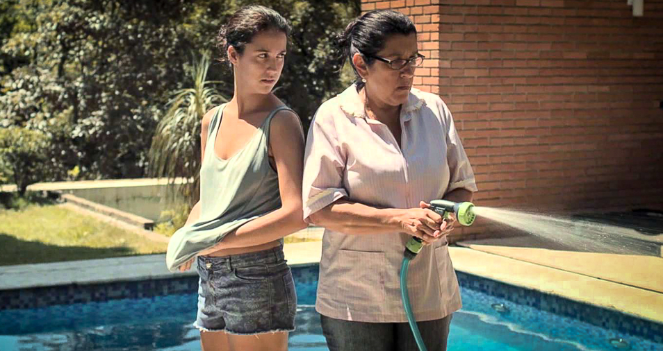Jéssica (Camila Márdila) and Val (Regina Casé) standing back to back next to the pool.